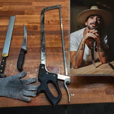 Background: a butcher block table with the tools of this craft on top, including two knives, a bone saw, and a cut-resistant glove. In the upper right hand corner, is an inset picture of Andrew Magazine with his chin resting on this hands and a broad brimmed hat on his head against a dark background.
